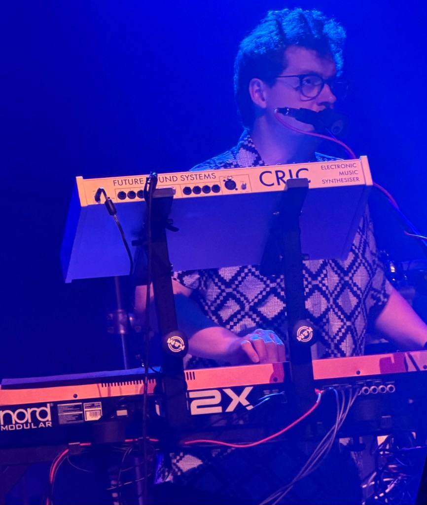 A musician playing an electronic synthesiser on stage, wearing glasses and a patterned shirt, with blue stage lighting in the background.