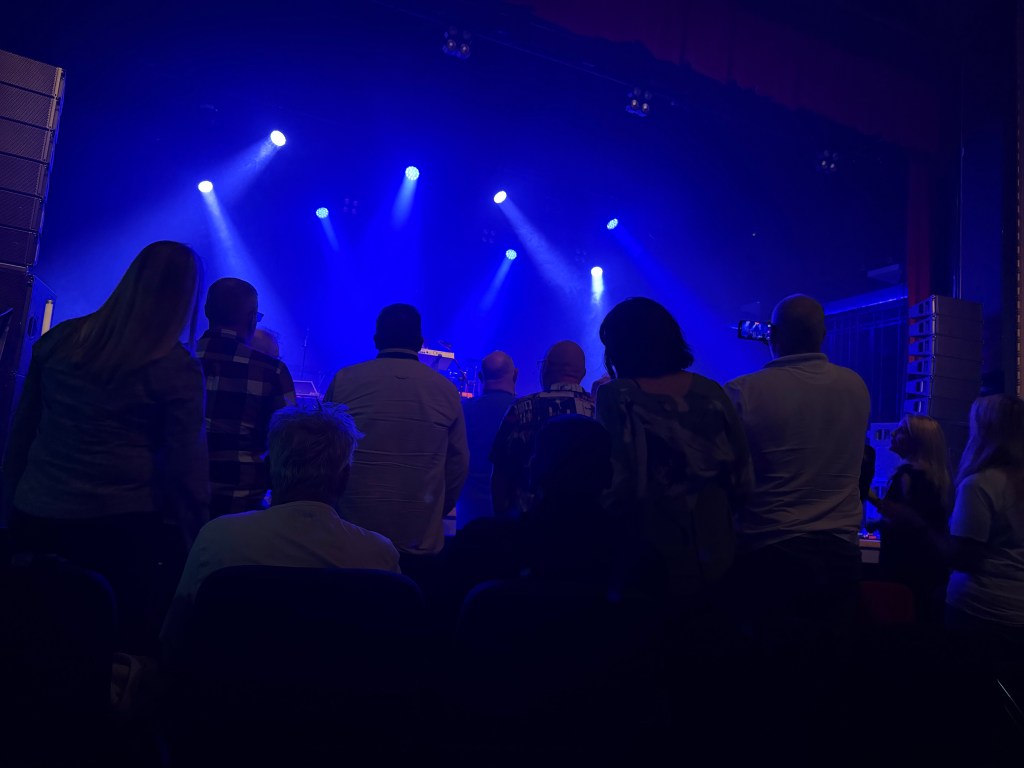 Audience members watching a performance in a dimly lit venue with blue stage lights.