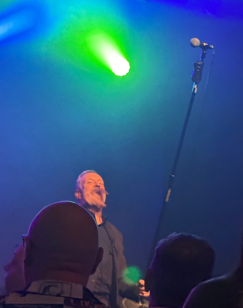 A performer singing passionately on stage with a microphone, surrounded by audience members in low light with colourful stage lighting.