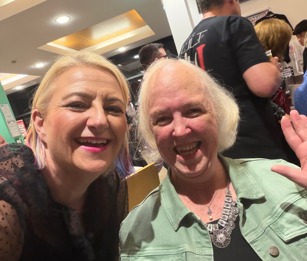 Two women smiling and posing for a selfie, one with blonde hair and a black top, the other with white hair wearing a mint green jacket and a silver necklace.