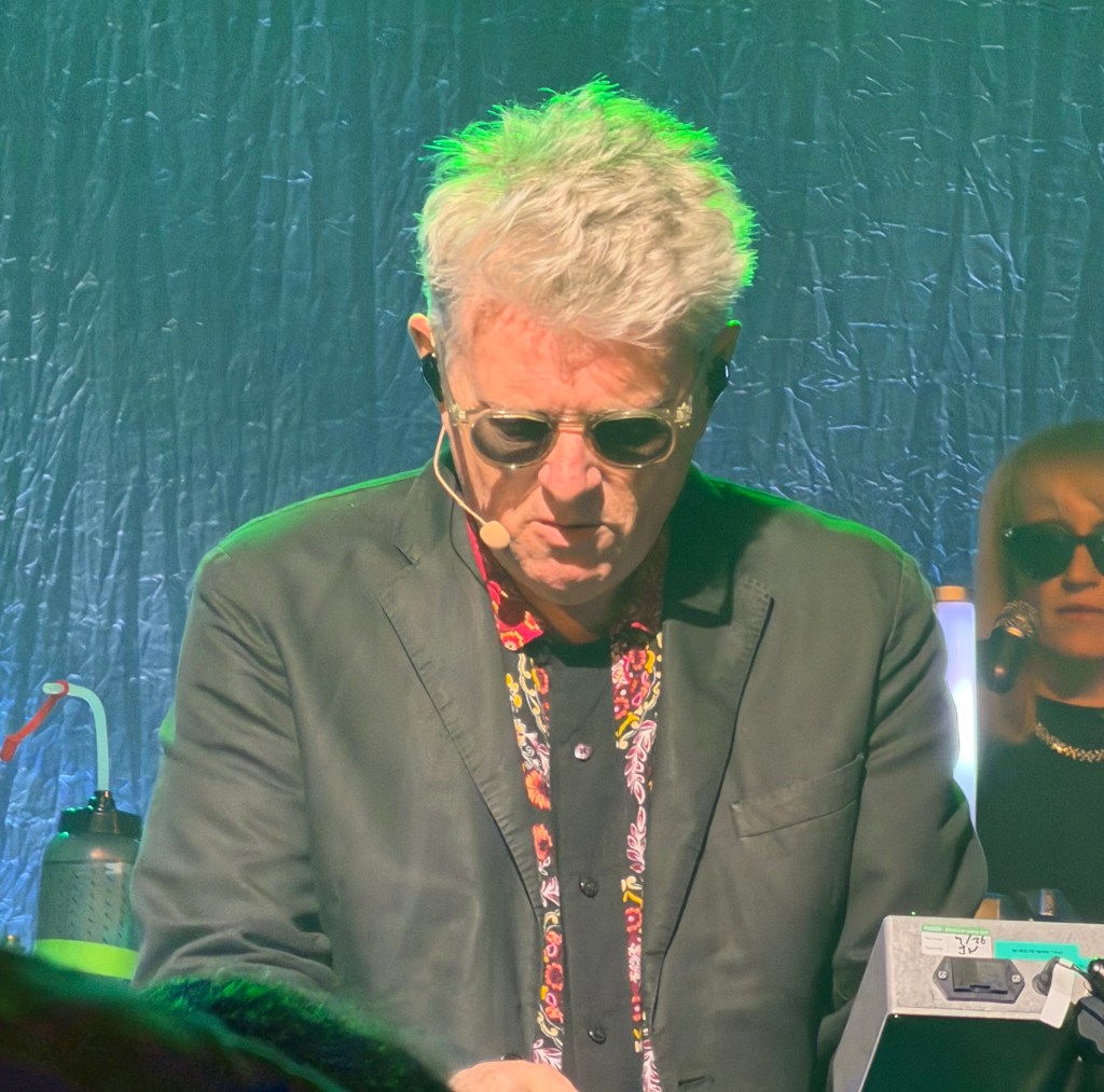 A musician wearing sunglasses and a black blazer, focused on performing with a microphone headset, adorned with a colourful floral shirt, in a vibrant, illuminated setting.