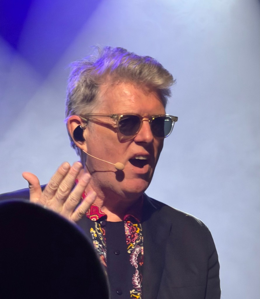 A man wearing sunglasses and a microphone passionately speaking on stage, with a stylish patterned shirt and a suited jacket, set against a hazy blue-lit background.