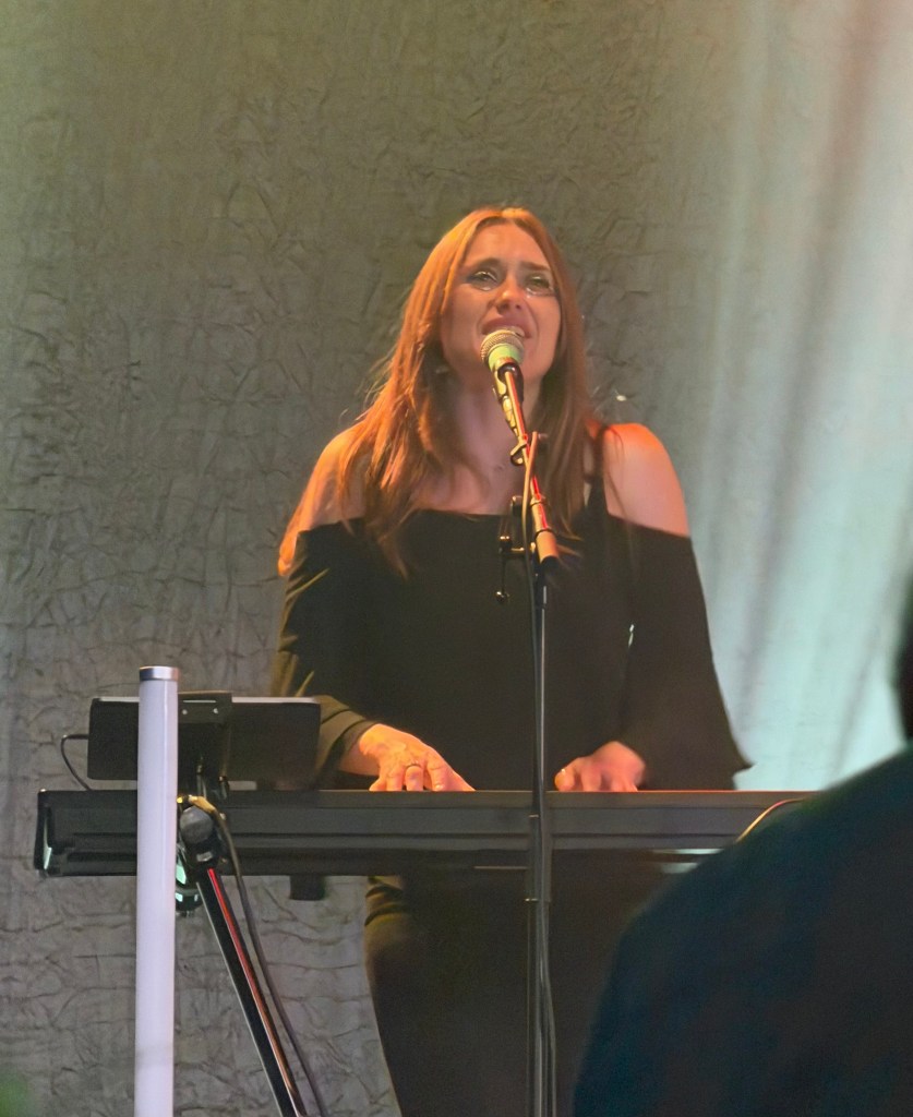 A female singer performing passionately at a keyboard, with emotional expression and a microphone in front of her, under soft stage lighting.