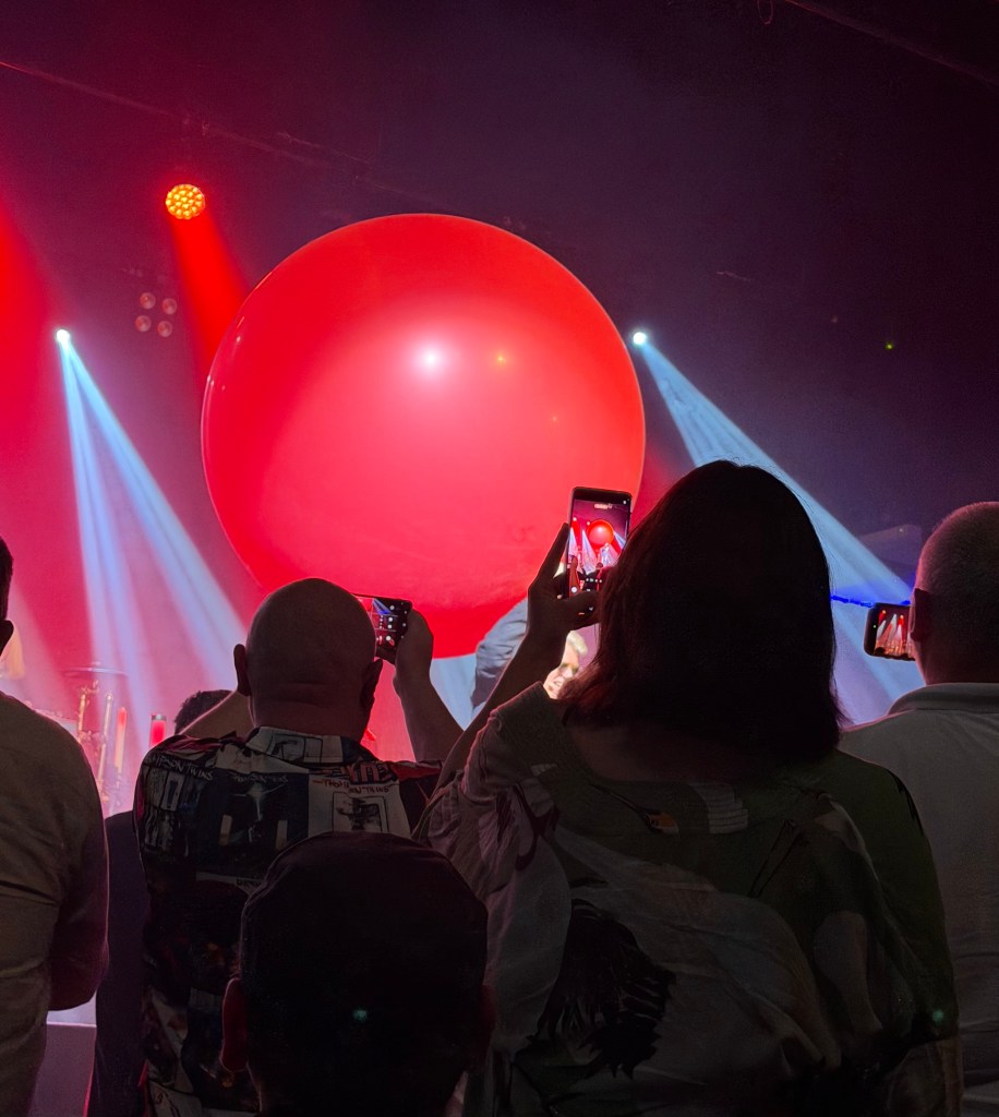 Audience members holding phones to capture a large red balloon on stage, with stage lights creating an atmospheric backdrop.