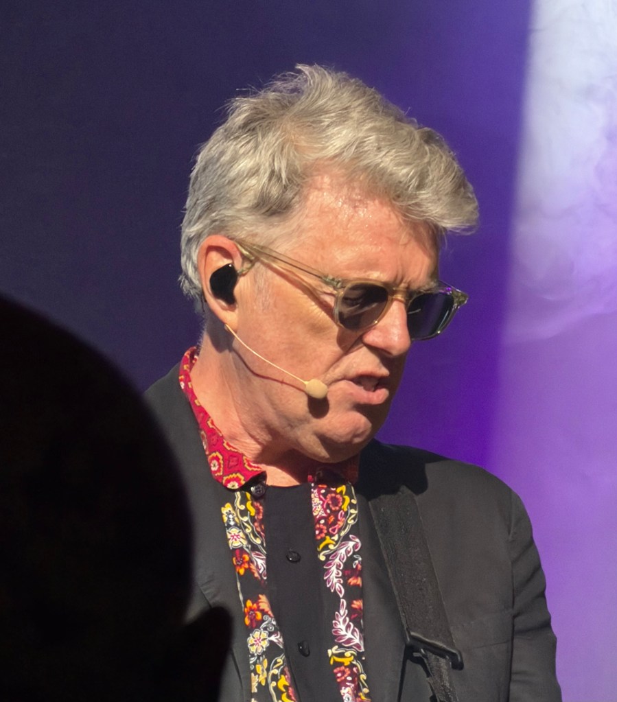 A musician with grey hair performing on stage, wearing sunglasses and a patterned shirt, with a microphone headset and guitar.