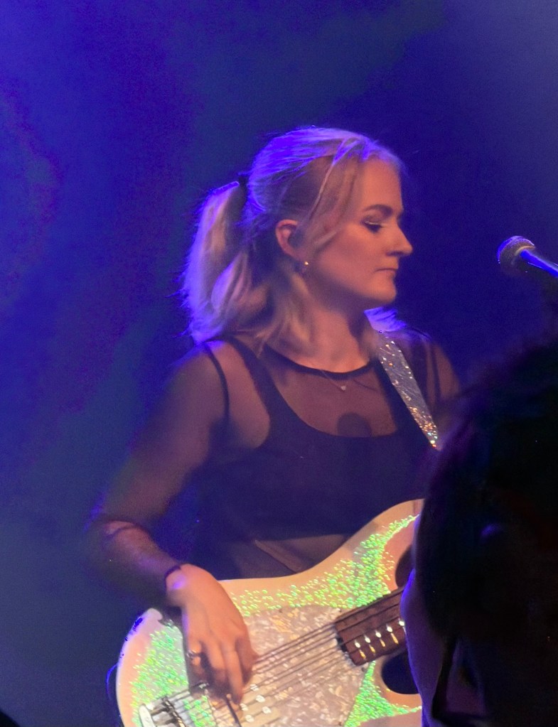 A female musician playing a glittery bass guitar on stage, illuminated by blue and purple lighting, with her hair pulled back.