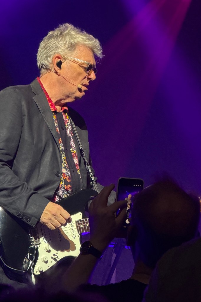 A musician performing on stage with a black electric guitar, wearing a stylish jacket and a patterned shirt, under purple stage lights. An audience member is capturing the moment with a smartphone.