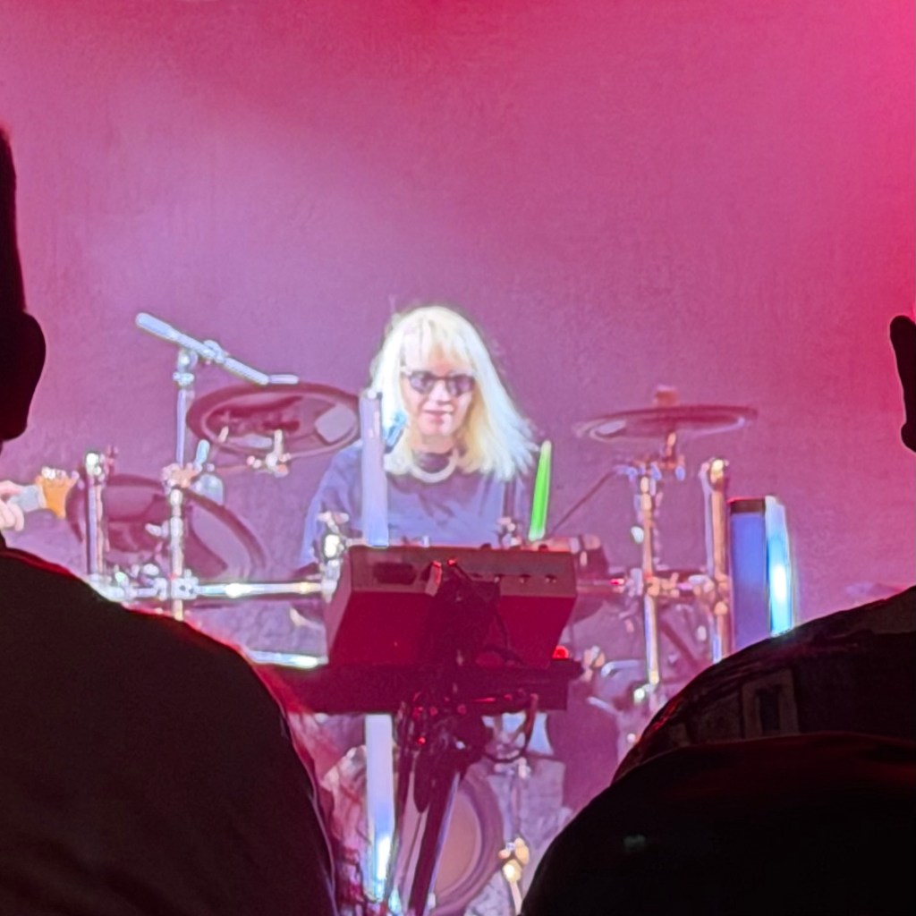 A performer with long blonde hair and sunglasses plays an electronic drum kit, with vibrant stage lighting in the background. Silhouettes of audience members are visible in the foreground.
