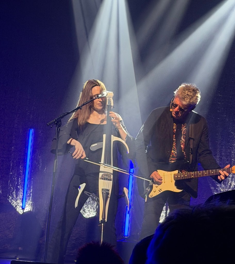 A female musician playing an electric cello while standing next to a male guitarist performing on stage, illuminated by dramatic lighting.